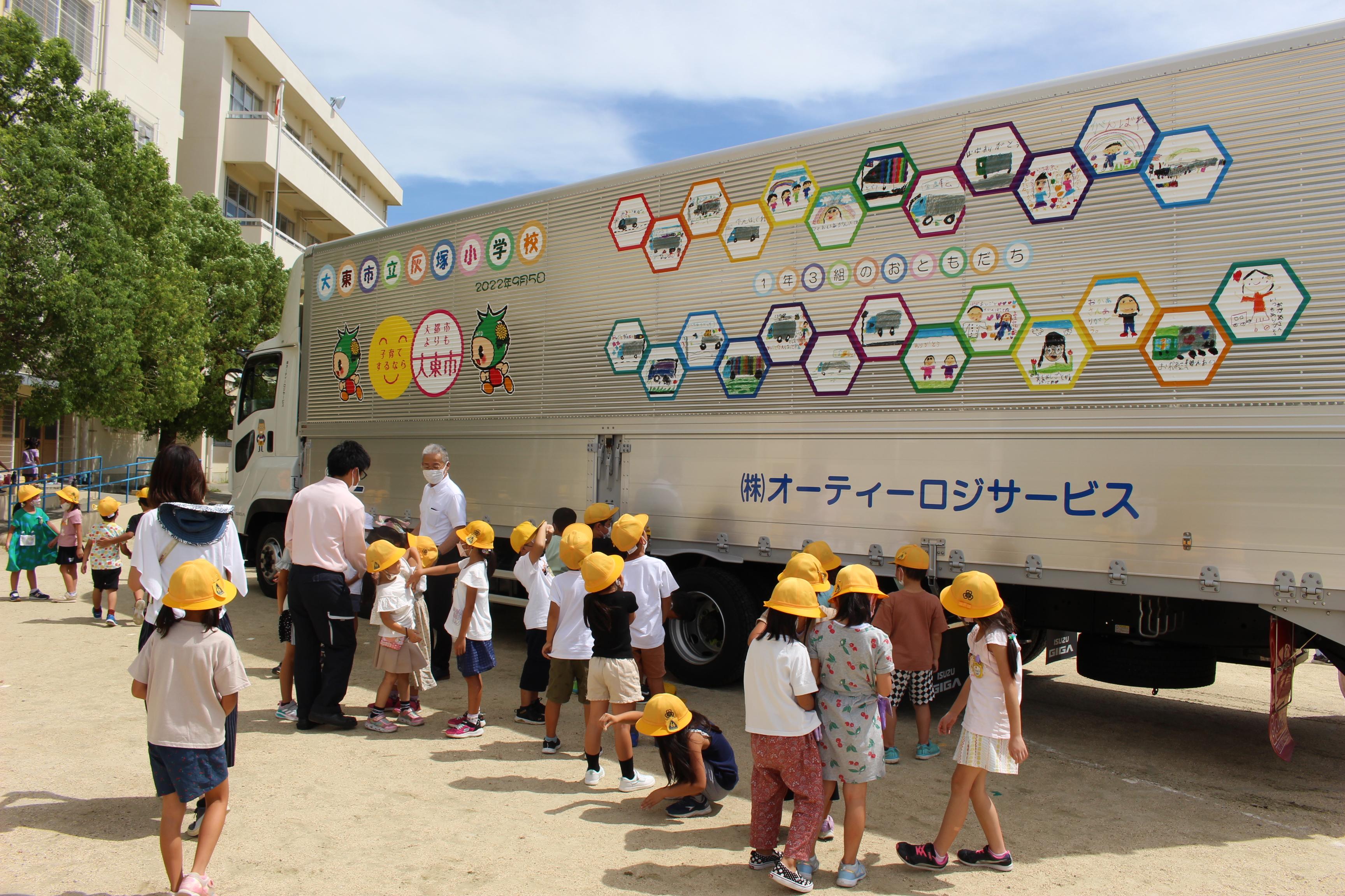 近所の小学校1年生の皆さんに描いてもらった絵をラッピングしたトラックの納車式典の様子です。